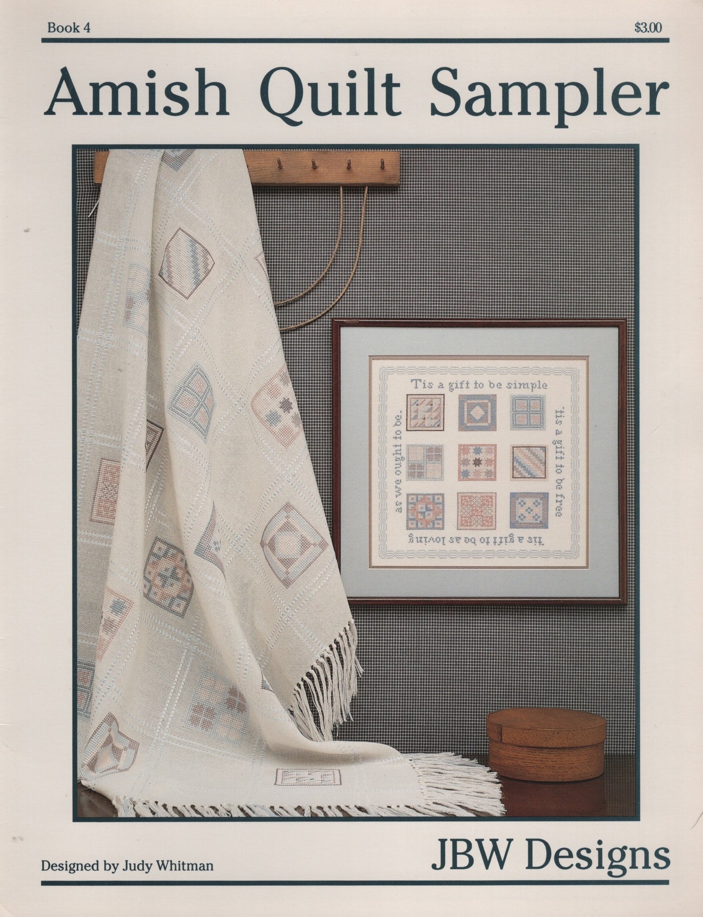 Cover of the Amish Quilt Sampler book by Judy Whitman, published by JBW Designs. The cover features a photograph of an off-white Amish quilt draped over a wall next to a framed cross stitch sampler design. Book 4