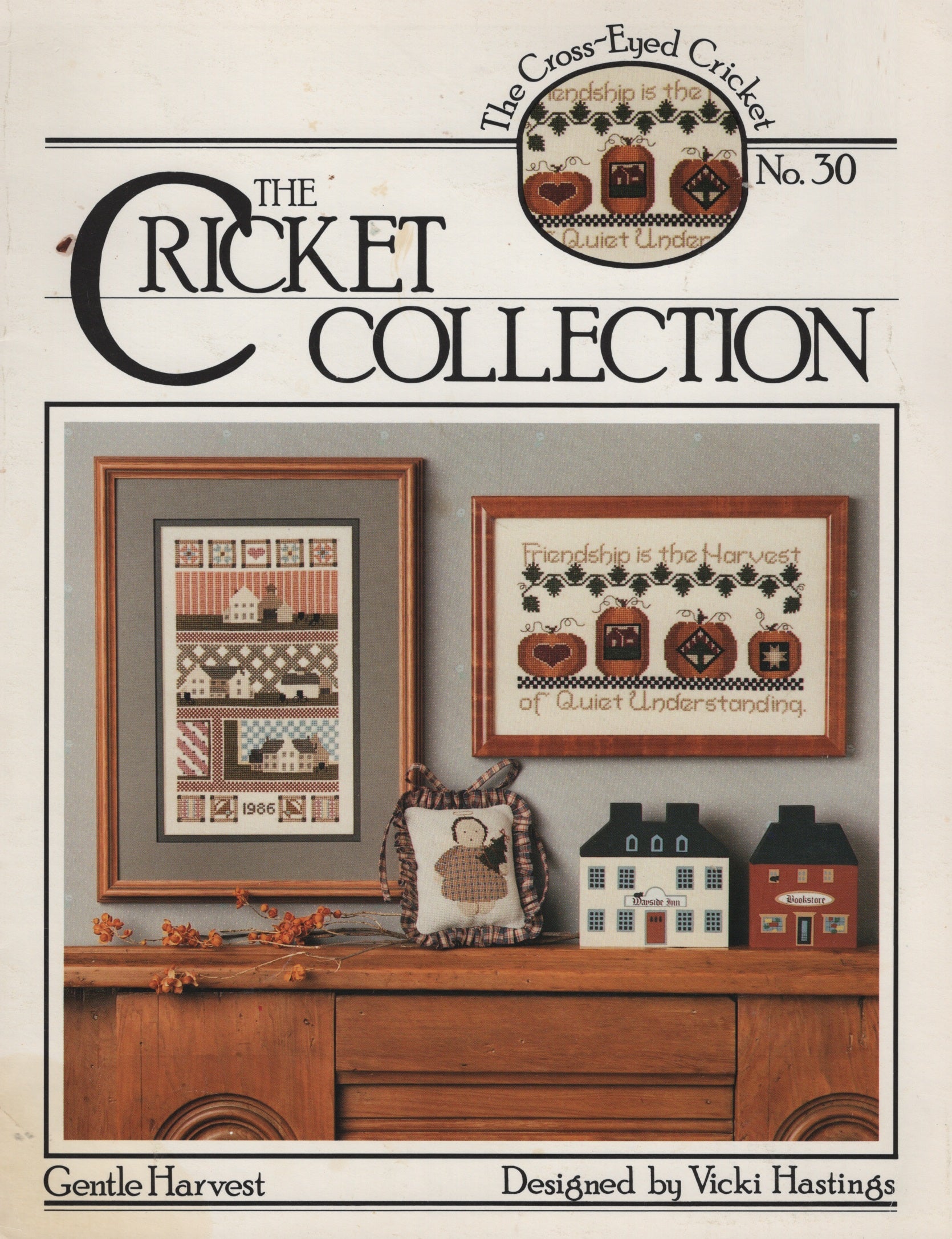 Front cover of The Cross-Eyed Cricket No. 30 'Gentle Harvest' pattern book by Vicki Hastings, showing framed cross-stitch designs and small house decorations on a wooden cabinet. CC30